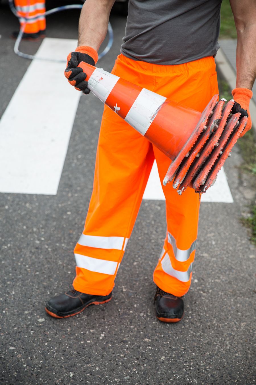 Worker in orange reflective trousers carrying a stack of traffic cones across a pedestrian crossing