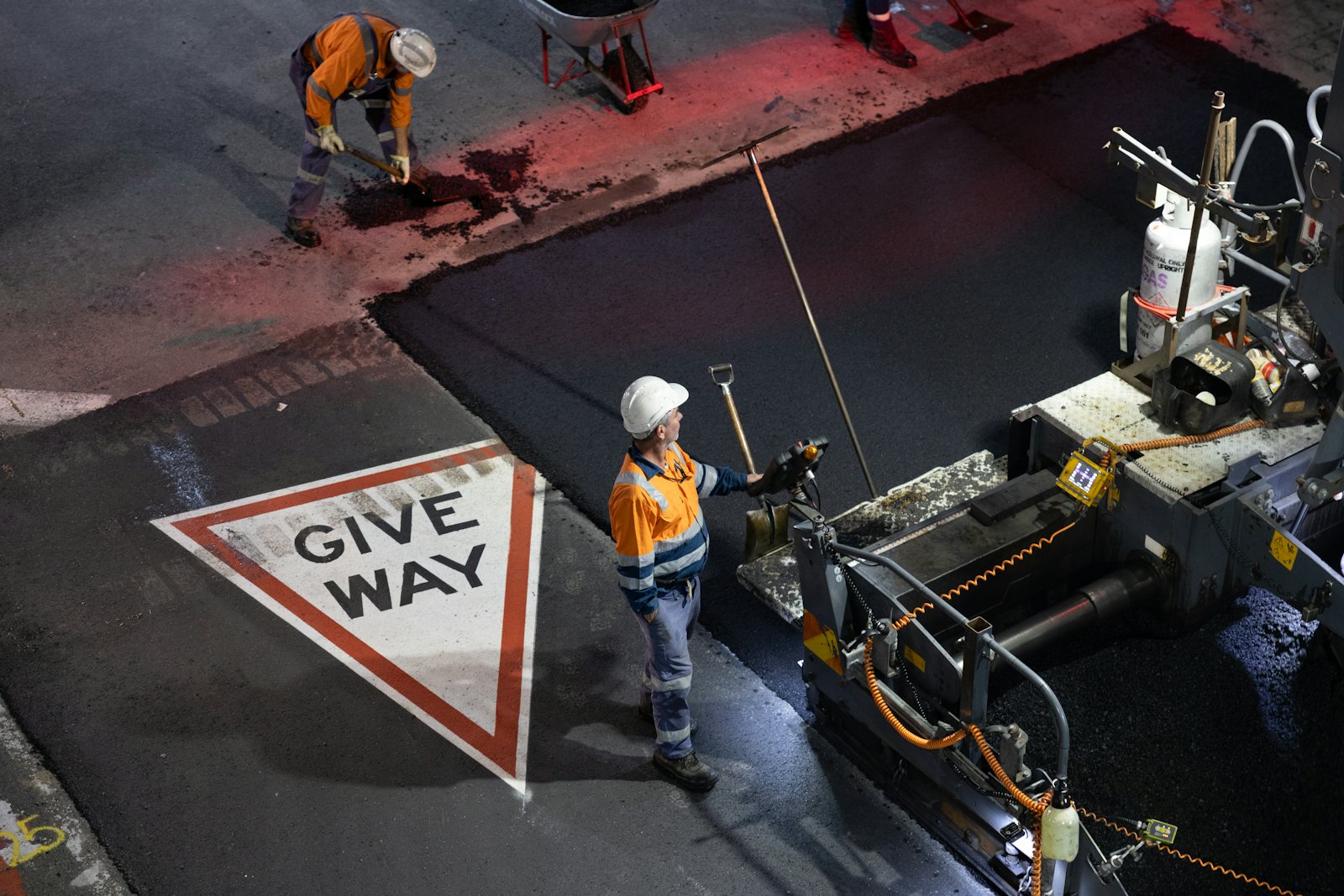 Road crew in high-visibility workwear resurfacing an intersection at night beside a painted Give Way marking